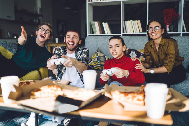 Amigos felices enfocados en ropa casual de moda jugando en la consola mientras se enfría en un sofá moderno y come pizza en un loft elegante - Foto, Imagen