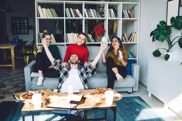 Friendly young gamers celebrating victory of man gladly raising hands up with game pad and eating pizza sitting on sofa in living room with book shelves  - Fotografie, Obrázek