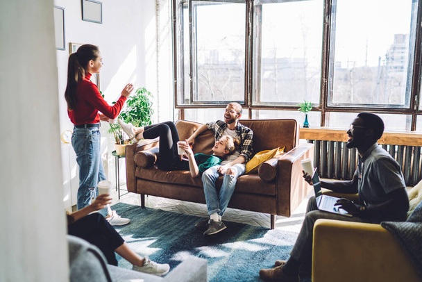Faceless young female in red long sleeve and jeans talking with multiethnic friends in high spirits taking comfortable places in bright room - 写真・画像