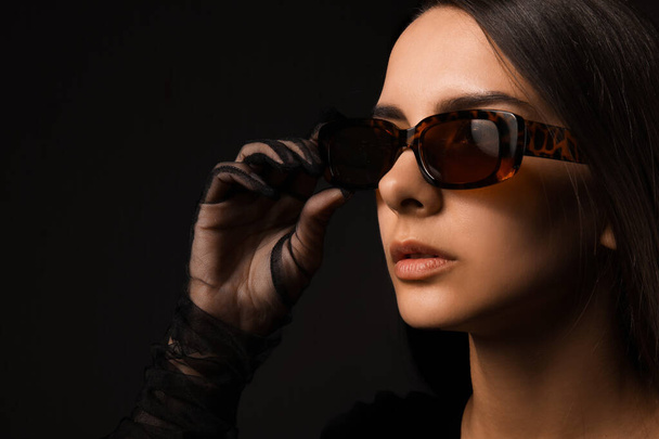 Young woman wearing stylish sunglasses on dark background - Foto, afbeelding