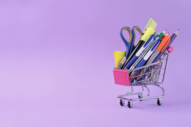 Metal shopping trolley with school supplies on a lilac background. School shopping concept. - Photo, Image