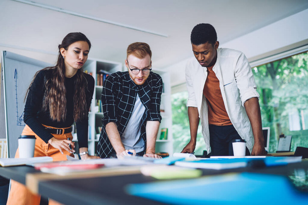 Multiracial young coworkers in casual clothes talking about data while standing around table and working on scheme during work in contemporary office - Foto, Bild