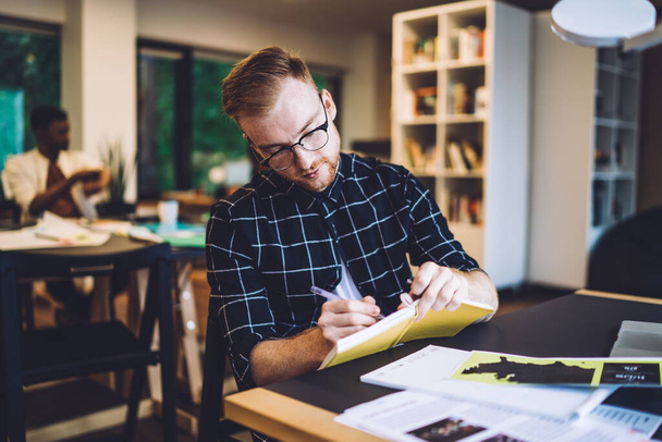 Concentrated male millennial making memo notes during learning knowledge project in coworking space for students, smart casual hipster guy writing test answer in paper textbook for education - Foto, Imagem