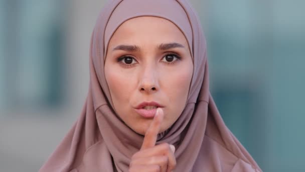 Primer plano serio retrato de la cara femenina al aire libre joven musulmana mujer islámica en hijab se para en la calle mirando la cámara sostiene el dedo índice cerca de la boca hace silencio gesto silenciar secretos prohíbe hablar - Imágenes, Vídeo