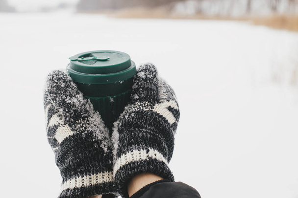 Ręce w przytulne rękawiczki gospodarstwa wielokrotnego użytku ciepłą filiżankę herbaty na tle jeziora śniegu w zimie. Plastikowy kubek z ciepłym napojem. Wędrówki i podróże w zimnym sezonie zimowym. Zrównoważony styl życia - Zdjęcie, obraz
