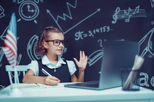 beautiful little schoolgirl sitting at desk and study online with laptop against black background with USA flag - Foto, Bild