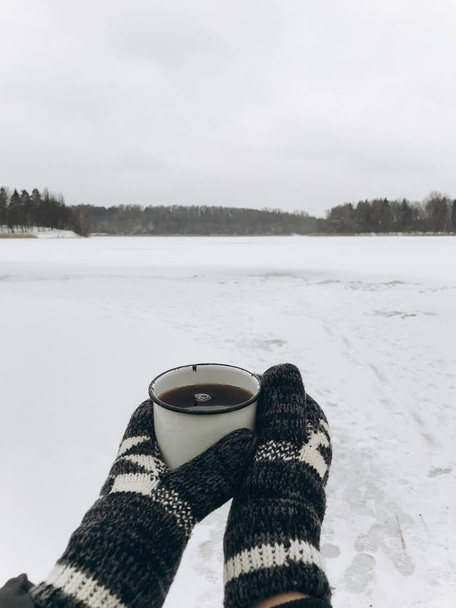 Ruce v útulných rukavicích s teplým šálkem čaje na pozadí sněžného jezera v zimě. Turistika a cestování v chladné zimní sezóně. Teplý drink. Prostor pro text. Toulavý - Fotografie, Obrázek