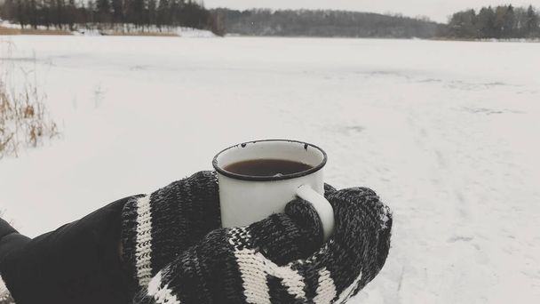 Hands in cozy gloves holding warm cup of tea on background of  snow lake in winter. Hiking and traveling in cold winter season. Warm drink. Space for text. Wanderlust - Photo, Image