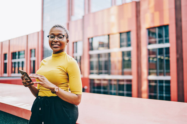 Half length portrait of cheerful woman in classic glasses for eyes correction holding cellphone technology and smiling at camera, African American female blogger with mobile gadget posing at urbanity - Photo, image