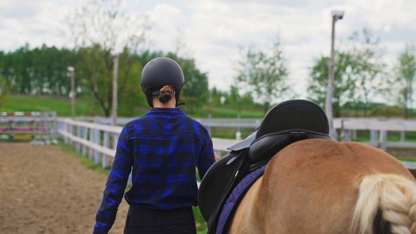 een jong jockey meisje leidt een paard naast een houten hek in de richting van obstakels - Foto, afbeelding