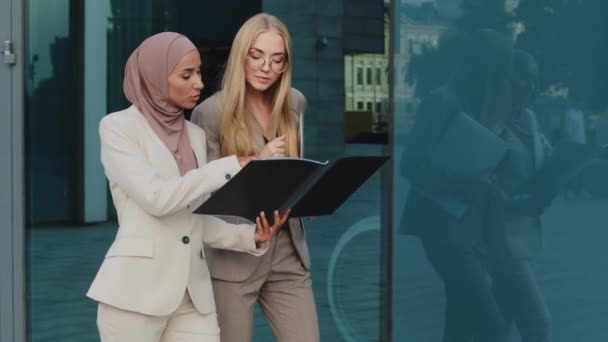 Duas mulheres de negócios diversas que andam ao longo da pasta de exploração de rua com documentos se comunicam antes da reunião, discutem o relatório. Menina indiana no hijab consulta com colega jovem mulher europeia durante o intervalo - Filmagem, Vídeo