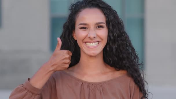 熱狂的な幸せな成功したブルネットの女の子ヒスパニック系の巻き女性が屋外の都市の建物の背景に立っている親指アップ同意のシンボル合意サポートサイン手ジェスチャー笑顔 - 映像、動画