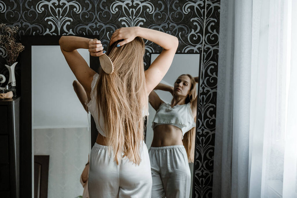 Comment prendre soin des cheveux longs. Vue arrière de la jeune femme blonde naturelle en vêtements de maison blanche peigner ses cheveux et regarder dans le miroir dans la chambre. - Photo, image