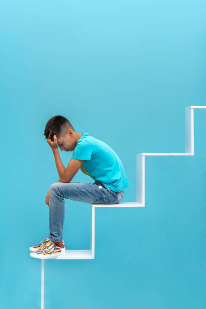 Plan en studio d'un jeune adolescent frustré ou fatigué portant une chemise bleue et un jean assis sur une échelle de développement faisant face à des difficultés sur un fond bleu. Concept d'éducation - Photo, image
