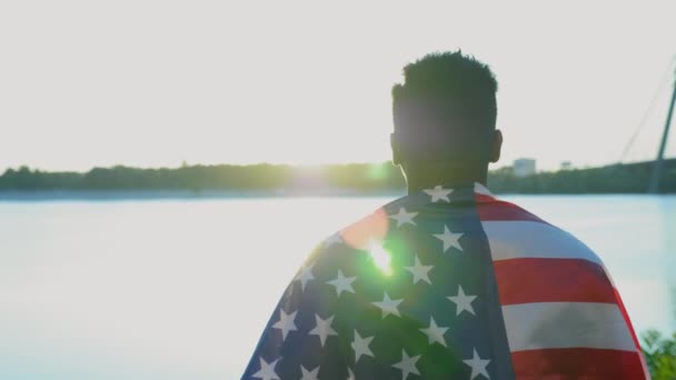 Αφρο-Αμερικανός άνδρας με αμερικανική σημαία στους ώμους κοιτάζει σε απόσταση κατά την ανατολή του ηλίου - Πλάνα, βίντεο