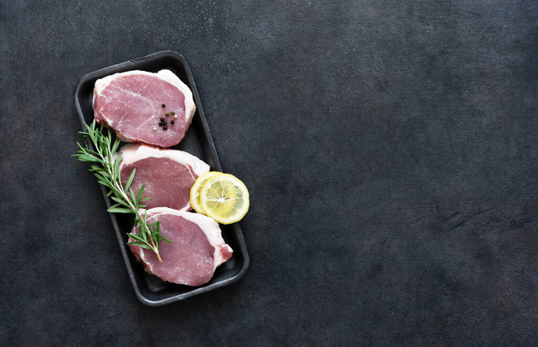 Raw pork steaks in a black tray with spices and lemon on a concrete background, top view. - Photo, Image