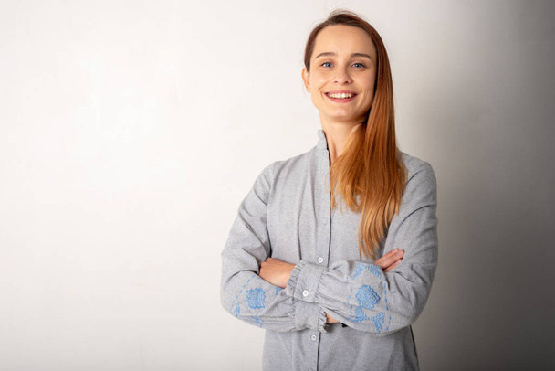 Confident woman in light gray clothes with blue eyes, blond hair and a smile on her face looks at the camera, standing by the white wall with her arms crossed. People, youth, beauty, lifestyle concept  - Photo, Image