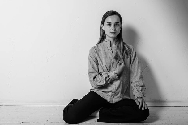 czarno-białe zdjęcie, młoda ładna dziewczyna siedząca na podłodze ze skrzyżowanymi nogami, wyglądająca prosto i na bok w szarej koszuli i czarnych spodniach, troskliwa, zdenerwowana, smutna w jasnym pokoju. Depresja i samotność - Zdjęcie, obraz
