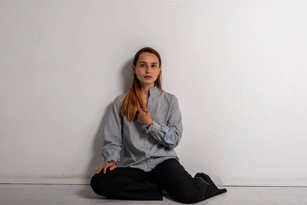 young pretty girl sitting on the floor crossed her legs looking straight at the camera in a gray shirt and black pants, thoughtful, upset, sad in a bright room. Depression and loneliness - Fotó, kép