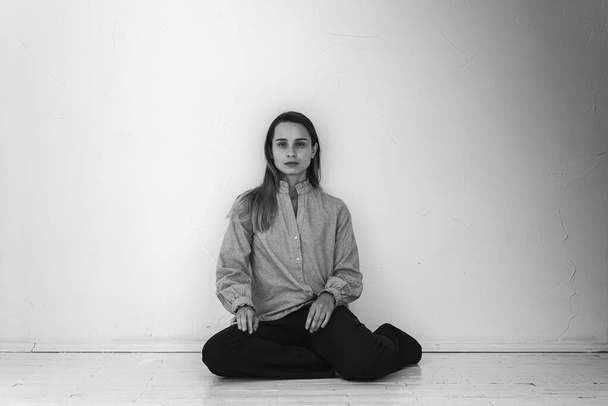 Schwarz-Weiß-Foto, ein junges hübsches Mädchen, das mit überkreuzten Beinen auf dem Boden sitzt und gerade und zur Seite schaut, in grauem Hemd und schwarzer Hose, nachdenklich, aufgebracht, traurig in einem hellen Raum. Depression und Einsamkeit - Foto, Bild