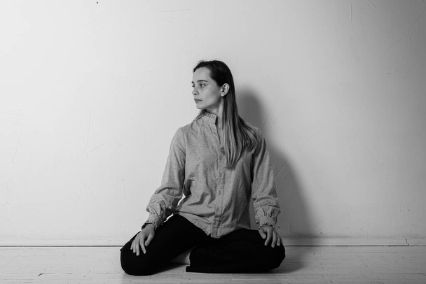 black and white photo, a young pretty girl sitting on the floor with her legs crossed looking straight and aside in a gray shirt and black pants, thoughtful, upset, sad in a bright room. Depression and loneliness - Photo, Image