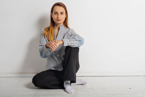 young pretty girl sitting on the floor crossed her legs looking straight at the camera in a gray shirt and black pants, thoughtful, upset, sad in a bright room. Depression and loneliness - Foto, Imagen