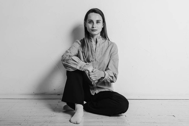 black and white photo, a young pretty girl sitting on the floor with her legs crossed looking straight and aside in a gray shirt and black pants, thoughtful, upset, sad in a bright room. Depression and loneliness - Photo, Image