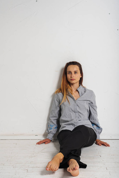 young pretty girl sitting on the floor crossed her legs looking straight at the camera in a gray shirt and black pants, thoughtful, upset, sad in a bright room. Depression and loneliness - Photo, Image