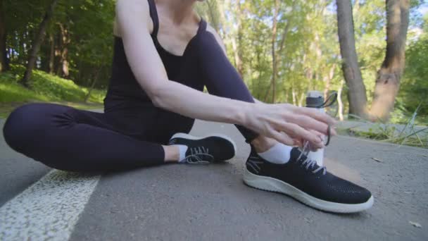Zbliżenie żeńskie nogi szczupłej biegaczki kobieta w czarnym stroju sportowym siedzi na torze w parku w pobliżu butelki wody po porannym joggingu prostuje sznurowadła na tenisówkach przygotowując się do biegania - Materiał filmowy, wideo