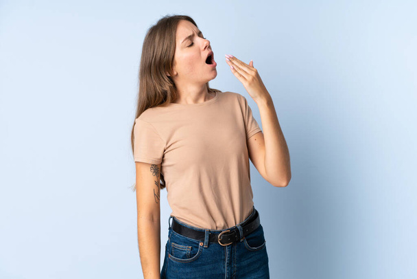 Young Lithuanian woman isolated on blue background yawning and covering wide open mouth with hand - Photo, Image