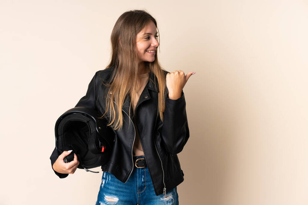 Mladá litevská žena držící motocyklovou přilbu izolované na béžovém pozadí ukazuje do strany prezentovat produkt - Fotografie, Obrázek