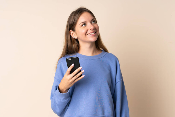 Junge Litauerin benutzt Handy isoliert auf beigem Hintergrund und schaut lächelnd nach oben - Foto, Bild