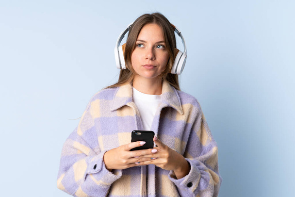 Jeune femme lituanienne isolée sur fond bleu écoutant de la musique avec un mobile et pensant - Photo, image