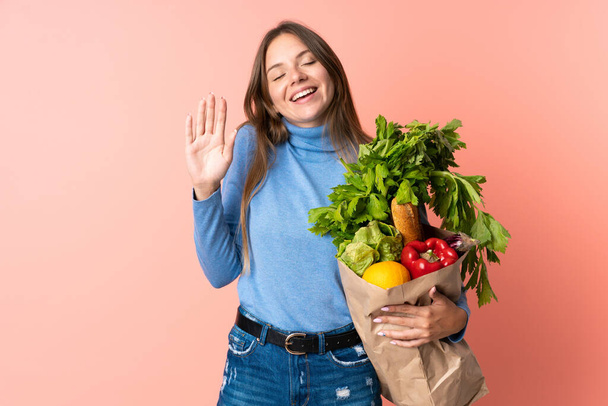 Young Lithuanian woman holding a grocery shopping bag saluting with hand with happy expression - Photo, Image