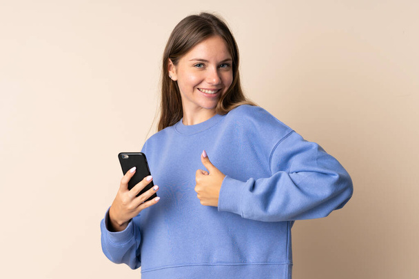 Junge Litauerin benutzt Handy isoliert auf beigem Hintergrund mit erhobenem Daumen, weil etwas Gutes passiert ist - Foto, Bild