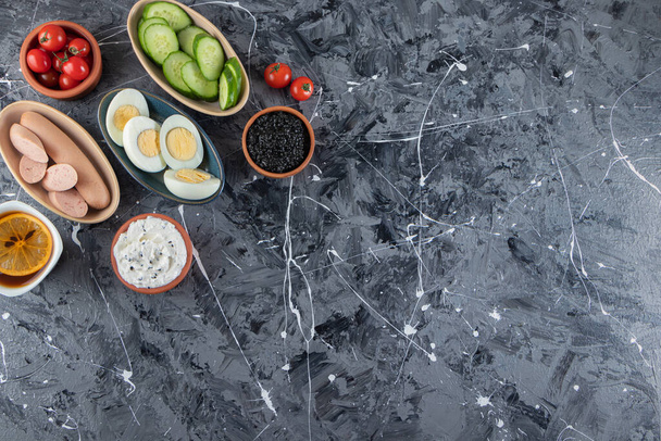 Sliced boiled eggs with fresh vegetables and boiled sausages placed on marble table  - Photo, Image