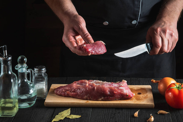 Ο σεφ κόβει κρέας με μαχαίρι στην κουζίνα, μαγειρεύει. Λαχανικά και μπαχαρικά στο τραπέζι της κουζίνας για να προετοιμάσει νόστιμο γεύμα - Φωτογραφία, εικόνα