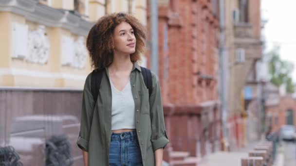 Fiatal, vegyes fajú, Y-generációs diáklány, göndör hajjal, alkalmi ruhában sétál a belvárosban. Vonzó afro-amerikai nő utazik egyedül a városban érzelmileg csodálja a modern építészet - Felvétel, videó