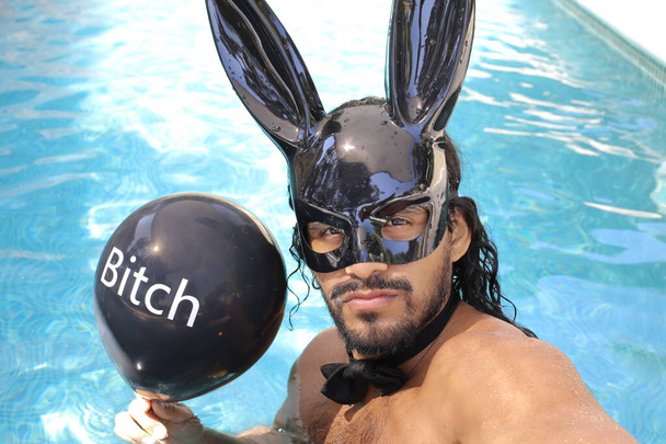 close-up portrait of handsome young mixed race shirtless man with bunny mask holding balloon with bitch inscription in pool - Photo, Image