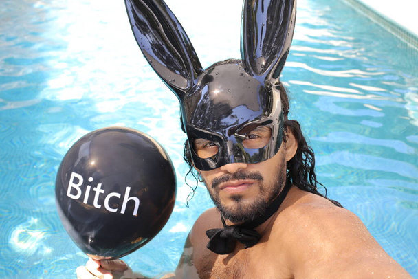 close-up portrait of handsome young mixed race shirtless man with bunny mask holding balloon with bitch inscription in pool - Photo, Image