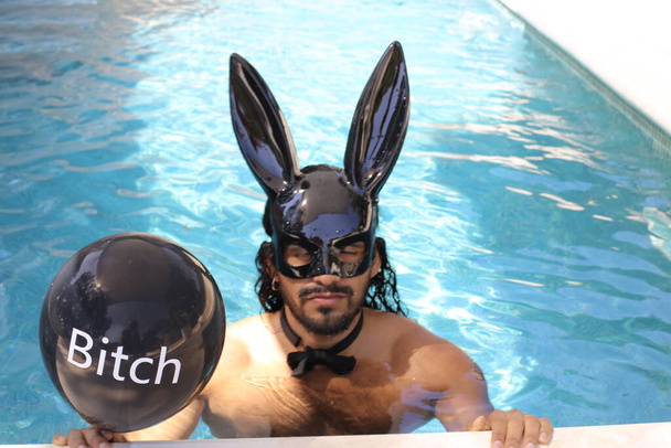close-up portrait of handsome young mixed race shirtless man with bunny mask holding balloon with bitch inscription in pool - Valokuva, kuva