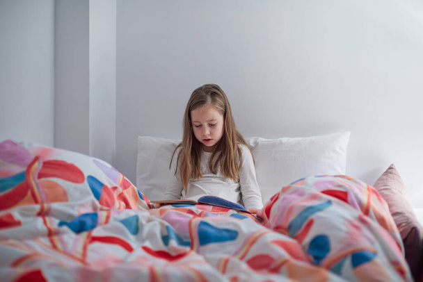 Yedi yaşında, yastığı ve büyük yorganıyla yatağında kitap ya da çizgi roman okuyan tatlı sarışın bir kız. - Fotoğraf, Görsel