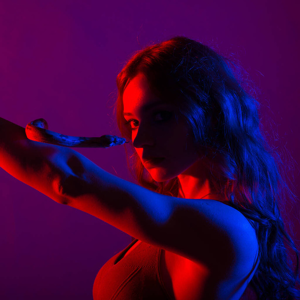 Όμορφη νεαρή κοκκινομάλλα γυναίκα με φίδι με ένα μοντέρνο τέλειο μακιγιάζ. Στιγμιότυπο στούντιο, νέον φως - Φωτογραφία, εικόνα