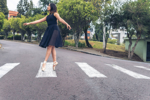 Bailarina latina adolescente con vestido negro caminando elegantemente con zapatos de ballet en un cruce peatonal en la calle - Foto, imagen