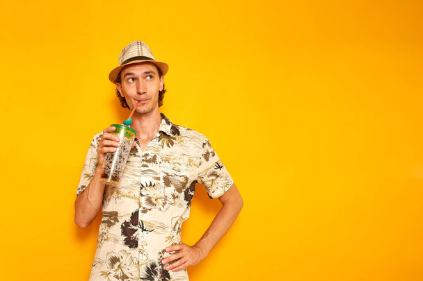 男性の旅行者を手に飲みながら休暇を過ごす旅行者は、麦藁を通してカクテルを楽しんだり、黄色の背景に隔離された帽子であるハワイのシャツの空のスペースに目を向けたりします。テキストのためのスペース。高品質の写真 - 写真・画像