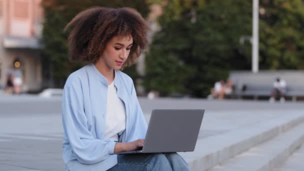 Afro american young girl user black mixed race ethnic business woman female student manager freelance worker sitting on sidewalk outdoors in city working with laptop e-learning online browsing website - Footage, Video