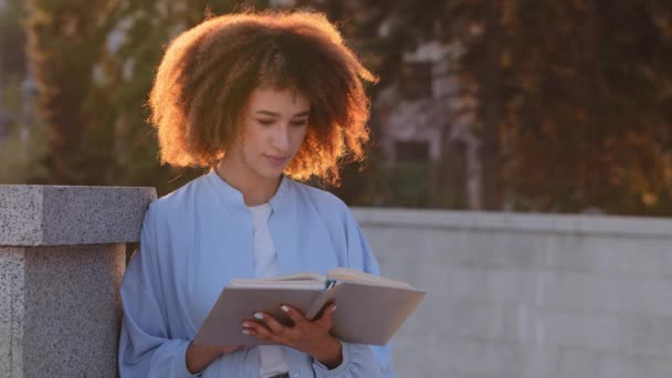 Afro amerykanka młoda afrykańska dziewczyna student czyta książki siedzi na ulicy w słońcu zachód słońca cieszy się literatura historia zamienia stronę czytanie informacji patrząc w notatnik - Materiał filmowy, wideo