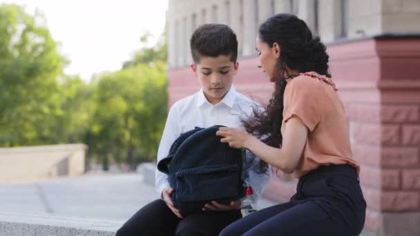 Mère hispanique femme avec un petit fils est assis à l'extérieur se préparant pour la classe scolaire parler. Maman aide écolier petit garçon élève élève avec la préparation pour la leçon met bouteille d'eau dans un cartable - Séquence, vidéo
