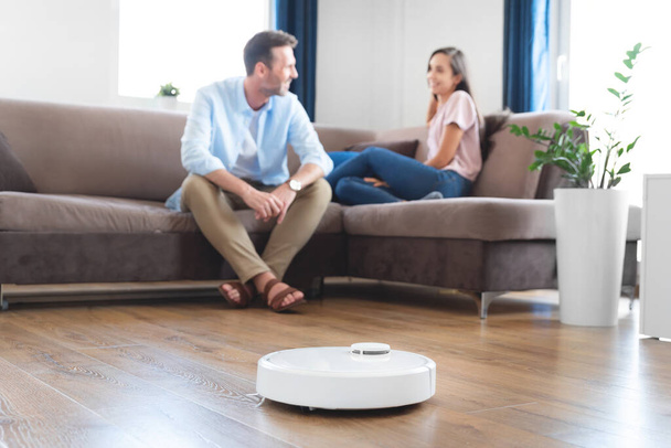 Robotic vacuum cleaner cleaning the room, smiling couple sitting on the sofa. Smart home concept - Foto, imagen