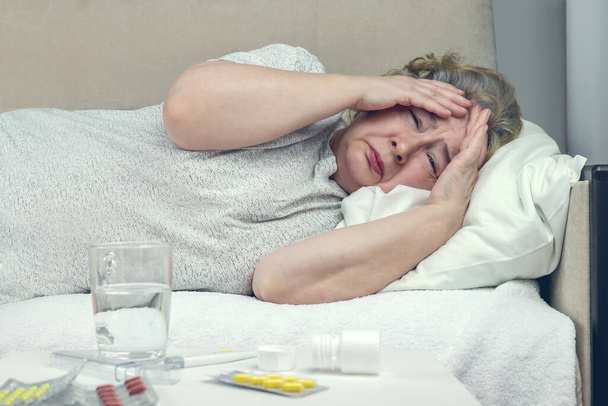 cierpiąca starsza kobieta leży w łóżku, jest chora trzymając głowę rękami, ma wysoką gorączkę i ból głowy - Zdjęcie, obraz
